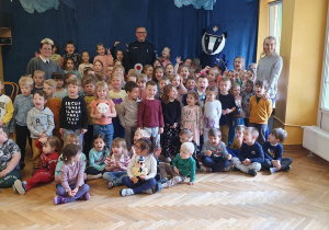 Dzieci siedzą i siedzą i pozują do wspólnego zdjęcia z wychowawczyniami, Panem policjantem oraz policyjną maskotką Borsukiem.
