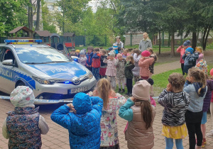 Dzieci stoją w ogródku przedszkolnym i słuchają dźwięku syreny policyjnej tzn. koguta.