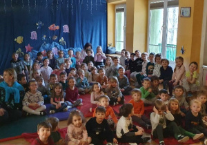 Dzieci z wszystkich grup siedzą na materacach oraz ławeczkach i czekają na rozpoczęcie się przedstawienia.