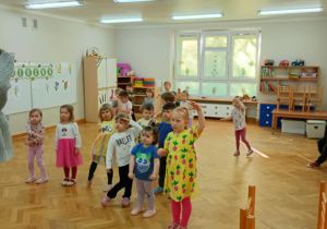 Dzieci stoją w rozsypce na sali i powtarzają taneczne ruchy za prowadzącą zajęcia z zumby, przykładając jedną rączkę do czoła.