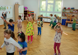 Dzieci stoją w rozsypce na sali i powtarzają taneczne ruchy za prowadzącą zajęcia z zumby, podnosząc rączki nad głowę.