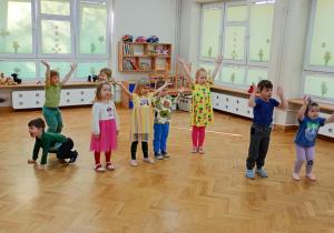 Dzieci stoją w rozsypce na sali i powtarzają taneczne ruchy za prowadzącą zajęcia z zumby, podnosząc obie rączki do góry w literkę V.