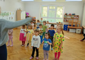 Dzieci stoją w rozsypce na sali i powtarzają taneczne ruchy za prowadzącą zajęcia z zumby.