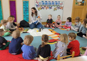 Dzieci siedzą na dywanie w półkolu i żegnają się z kukiełką Profesorka Labolka i prowadzącą.