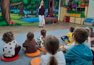 Technoludek stoi na środku sali, za nim rozciągnięte na stelażu wisi tło lasu.