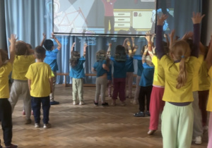 Dzieci ubrane w niebieskie i żółte koszulki stoją przed tablicą i wykonują ćwiczenia na niej wyświetlane, unosząc wysoko ręce.