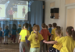 Dzieci ubrane w niebieskie i żółte koszulki stoją przed tablicą i wykonują ćwiczenia na niej wyświetlane, podnosząc nogę wysoko.