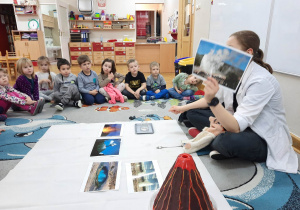 Dzieci siedzą na dywanie i oglądają prezentowane przez prowadząca warsztaty zdjęcia erupcji wulkanów.