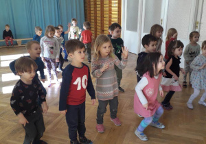 Dzieci stoją w rozsypce na sali gimnastycznej poruszają się w dowolny sposób do muzyki.