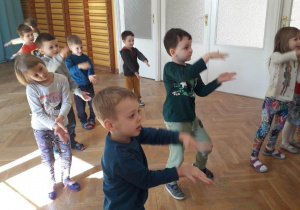 Dzieci stoją w rozsypce na sali gimnastycznej tańczą do piosenki z pokazywaniem klaszcząc w dłonie.