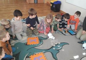 Dzieci siedzą na dywanie i próbują zaprogramować trasę poruszania się robocika za pomocą tabletu.