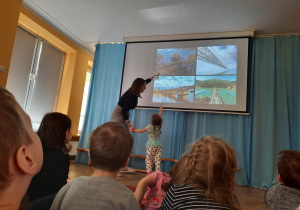 Dzieci siedzą na materacach i oglądają prezentację o mostach, a dziewczynka z prowadząca warsztaty wskazuje na jedno ze zdjęć.