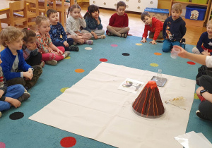 Dzieci siedzą na dywanie i obserwują jak prowadząca warsztaty LABOLO wykonuje eksperyment polegający na wlaniu płynu do wulkanu.