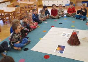 Dzieci siedzą na dywanie, prowadząca pokazuje im zdjęcia przedstawiające wulkan i jego erupcję.