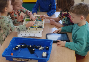 Dzieci siedzą przy stole i układają robota z klocków LEGO na podstawie instrukcji wyświetlanej na tablecie na zajęciach z robotyki.