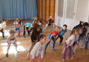 Dzieci stoją w rozsypce na sali gimnastycznej powtarzają ruchy taneczne za trenerką zumby stojąc w rozkroku z lekko ugiętymi kolanami, pochylając się do przodu i opierając ręce na kolanach.
