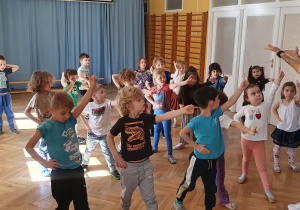 Dzieci stoją w rozsypce na sali gimnastycznej powtarzają ruchy taneczne za trenerką zumby wyciągając rękę do przodu, a drugą trzymają na biodrach.