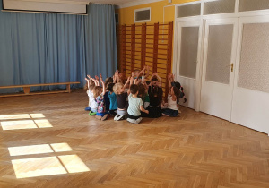 Dzieci siedzą w rogu sali z rękami uniesionymi do góry.