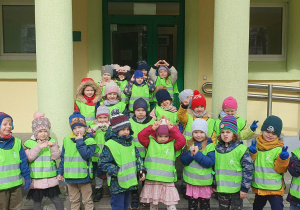 Dzieci stoją przed wejściem do przedszkola ubrane w kamizelki odblaskowe pozują do zdjęcia przed wyjściem na wycieczkę.