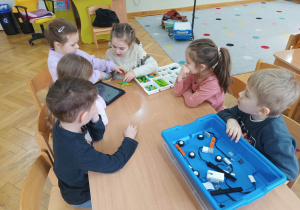 Dzieci siedzą przy stoliku i układają robota z klocków LEGO na podstawie instrukcji wyświetlanej na tablecie na warsztatach z robotyki.