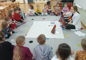 Dzieci siedzą na dywanie i zapoznają się z wewnętrzną budową wulkanu przedstawioną na modelu.