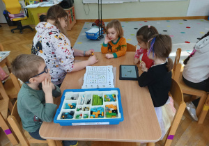 Dzieci siedzą przy stoliku i z pomocą prowadzącej zajęcia układają robota z klocków LEGO na podstawie instrukcji wyświetlanej na tablecie na warsztatach z robotyki.
