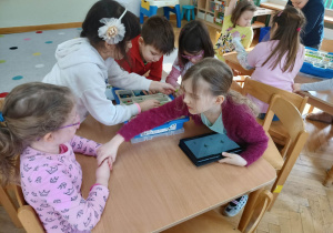 Dzieci siedzą przy stolikach i układają robota z klocków LEGO na podstawie instrukcji wyświetlanej na tablecie na warsztatach z robotyki.