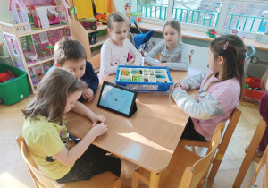Dzieci siedzą przy stoliku i układają robota z klocków LEGO na podstawie instrukcji wyświetlanej na tablecie na warsztatach z robotyki.