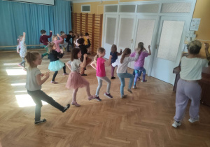 Dzieci stoją ustawione na sali gimnastycznej i powtarzają kroki taneczne za trenerką zumby wykonując wymach ręką i nogą.