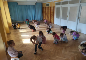 Dzieci stoją ustawione na sali gimnastycznej i powtarzają kroki taneczne za trenerką zumby kucając i chowając głowę między rękami.