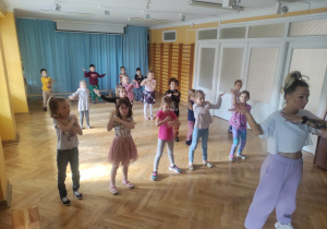 Dzieci stoją ustawione na sali gimnastycznej i powtarzają kroki taneczne za trenerką zumby kładąc ręce na ramionach.