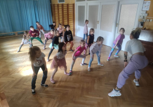 Dzieci stoją ustawione na sali gimnastycznej i powtarzają kroki taneczne za trenerką zumby przykucając z szeroko rozstawionymi nogami.