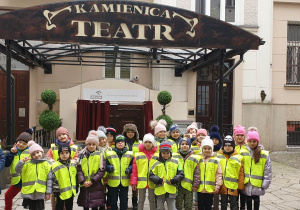 Dzieci stoją przed wejściem do Teatru Kamienica ubrane w kamizelki odblaskowe pozują do zdjęcia przed spektaklem.