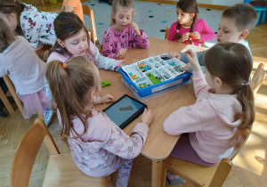 Dzieci siedzą przy stoliku i za pomocą instrukcji wyświetlanej na tablecie budują roboty z klocków LEGo na warsztatach z robotyki i programowania.