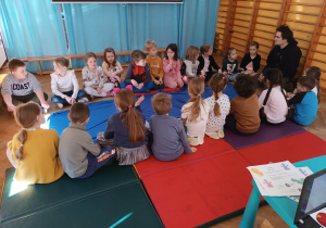 Dzieci siedzą po dwóch stronach niebieskiego materiału i rozmawiają z prowadzącym warsztaty o rzece Wiśle.
