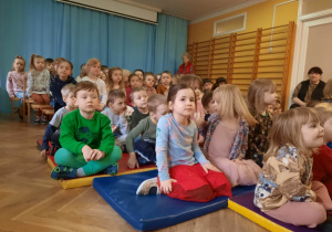 Dzieci siedzą na ławkach oraz materacach i oglądają przedstawienie wystawiane w przedszkolu na sali gimnastycznej.