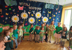 Dzieci ubrane na zielono stoją w półkolu i śpiewają piosenkę przygotowaną dla Pani Wiosny na powitanie.