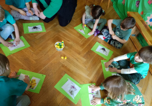 Dzieci siedzą w kole na podłodze i układają puzzle z pociętych na kawałki obrazków wiosennych kwiatków.