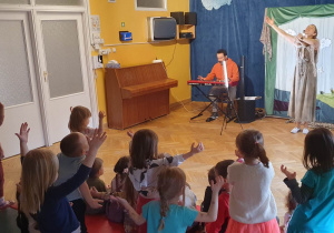Dzieci powtarzają ruchy za Panią prowadzącą koncert machając wyprostowanymi rękami na boki, a Pan gra na keyboardzie.