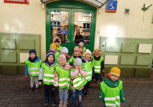Dzieci stoją przed wejściem do przedszkola ubrane w kamizelki odblaskowe gotowe na wycieczkę.