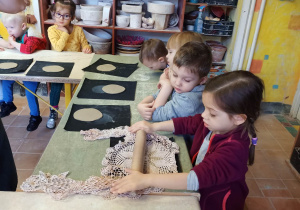 Dzieci siedzą przy stole, a dziewczynka wałkuje placek z gliny odciskając wzorem z serwetki na nim.