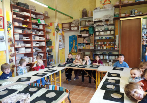 Dzieci siedzą przy stolikach i prezentują wykonane przez siebie talerzyki z gliny na warsztatach ceramicznych w pracowni ceramicznej Amfora.