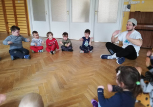 Dzieci siedzą na podłodze na sali gimnastycznej i powtarzają ćwiczenia za prowadzącym zajęcia sportowe.