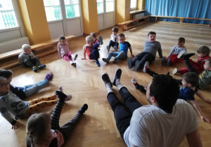 Dzieci siedzą na podłodze na sali gimnastycznej i powtarzają ćwiczenia za prowadzącym zajęcia sportowe, podnosząc na zmianę raz jedną, raz drugą wyprostowaną nogę..
