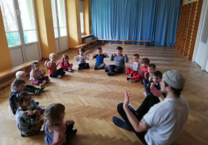 Dzieci siedzą na podłodze na sali gimnastycznej i powtarzają ćwiczenia za prowadzącym zajęcia sportowe.