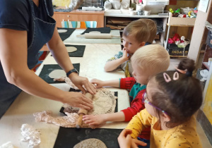 Dzieci siedzą przy stole, a chłopiec z pomocą prowadzącej warsztaty wałkuje placek z gliny odciskając wzorem z serwetki na nim.