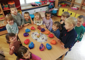 Dzieci stoją dookoła stołu i prezentują własnoręcznie pomalowane i ozdobione cekinami styropianowe zajączki wielkanocne.