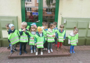 Dzieci stoją przed wejściem do przedszkola ubrane w kamizelki odblaskowe gotowe na wycieczkę.