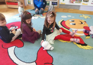 Dzieci siedzą na dywanie i witają się z robocikiem na zajęciach z robotyki i programowania.
