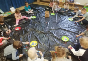 Dzieci siedzą w kole na folii i układają własne obrazy abstrakcyjne z leżących przed nimi kolorowych karteczek w różnych kształtach.